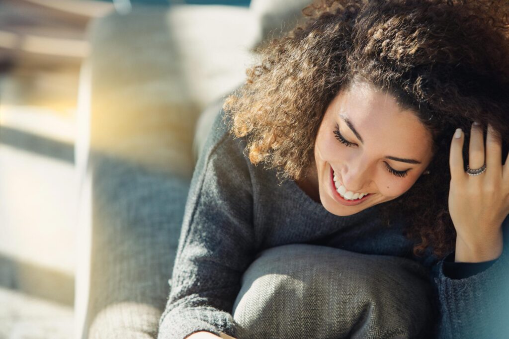 Young woman with curly hair sits happily on a cozy sofa, enjoying a peaceful moment at home. Sunlight adds tranquility, showcasing her joy and positivity in a natural setting